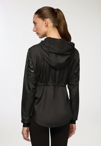 Czarna kurtka z kapturem, ściągnięta w talii, z błyszczącymi, wzorzystymi akcentami na rękawach; wykonana z lekkiego, gładkiego materiału.