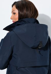 Mujer de cabello oscuro con una chaqueta con capucha azul marino, girada ligeramente hacia un lado sobre un fondo blanco.