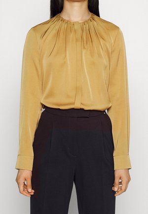 Femme portant un chemisier jaune moutarde à manches longues avec des plis au niveau de l'encolure, rentrée dans un pantalon noir taille haute.