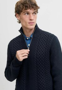Sudadera navy con cremallera y un patrón de punto texturizado, con puños y collar acanalados, superpuesta sobre una camisa de denim con cierre de botones.