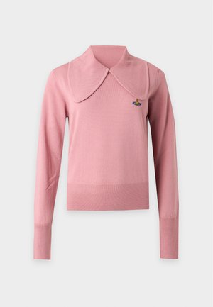 Femme portant un haut rose à manches longues, un pantalon beige à jambes larges, des chaussures noires pointues, tenant un sac à bandoulière noir texturé.