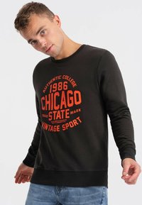 Felpa nera con grafica rossa che riporta il testo "1986 CHICAGO STATE VINTAGE SPORT." Presenta un collo tondo e polsini a costine. Indossata con jeans blu.