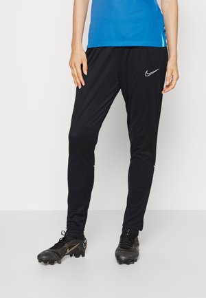 Person trägt schwarze Nike-Trainingshose, schwarze Fußballschuhe mit goldenen Akzenten und ein blaues Shirt vor einfarbigem Hintergrund.