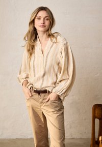 Femme aux longs cheveux blonds portant un chemisier ample à rayures crème et un pantalon beige, debout les mains dans les poches contre un mur neutre.