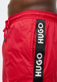 Piros rövidnadrág könnyű anyagból, elasztikus derékpánttal, fekete oldalsávval, amelyen a „HUGO” márkanév olvasható.