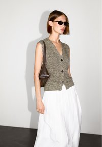 Eine Frau trägt schwarze Sonnenbrille, einen ärmellosen Strickpollover, einen weißen gefältelten Rock und hält eine dunkelbraune Umhängetasche, während sie vor einem einfarbigen Hintergrund steht.