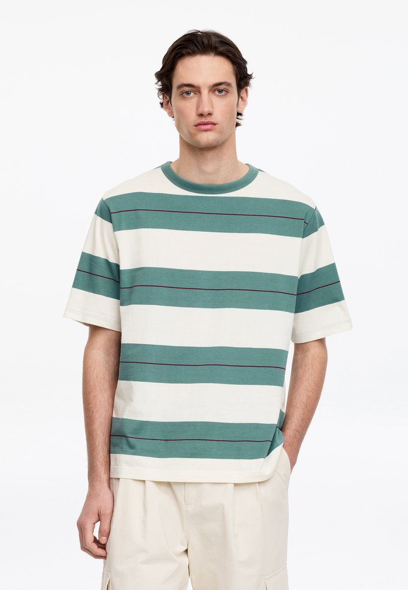 Jeune homme portant un t-shirt à manches courtes rayé turquoise et blanc avec un pantalon couleur crème, debout devant un fond blanc uni.