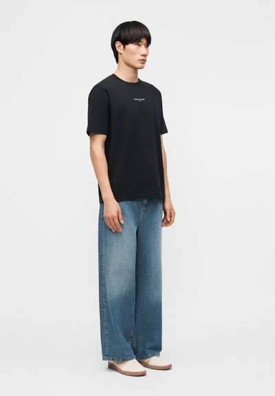 Άνδρας που στέκεται φορώντας μαύρο κοντομάνικο μπλουζάκι με λογότυπο, φαρδιά μπλε τζιν παντελόνια και λευκά παπούτσια σε απλό λευκό φόντο.