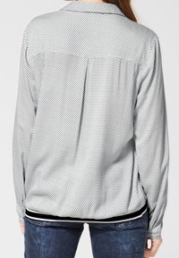 Blouse à manches longues avec col blanc à pois noirs, dotée d'un empiècement au dos et d'un ourlet rayé noir. Tissu lisse avec une texture douce.