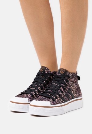 Beine in hoch geschnittenen Adidas-Sneakers mit pink-schwarz leopardenmuster, schwarzen Schnürsenkeln, weißen Sohlen und braunem Rand.