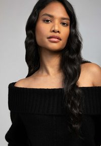 Pull en maille noir à épaules dénudées avec texture côtelée et large décolleté. Le mannequin a de longs cheveux sombres ondulés et un maquillage neutre.