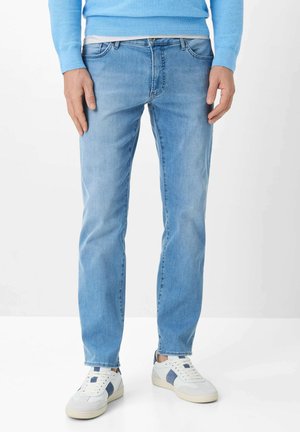 Man draagt lichtblauwe spijkerbroek, een blauwe trui, witte sneakers met blauwe accenten, en staat voor een effen witte achtergrond.