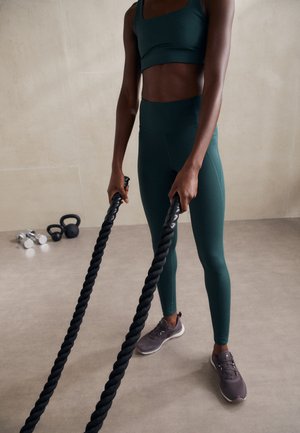 Athletic outfit in dark green fabric; sports bra and high-waisted leggings. Black battle ropes held in hands, gray sneakers on feet.