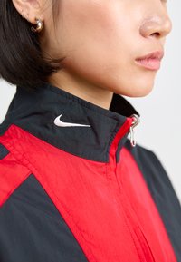 Nike vetrovka z visokim ovratnikom, črne in rdeče barvne kombinacije, srebrnim zadrgo in logotipom Nike na ovratniku.