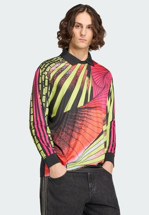 Joven con cabello hasta los hombros que lleva una camisa de manga larga colorida con patrones geométricos y jeans negros, con las manos en los bolsillos.