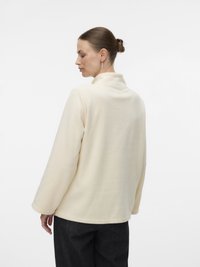 Γυναίκα με μαλλιά σε κότσο που φοράει κρέμα φλις πανωφόρι και μαύρο παντελόνι, απεικονιζόμενη από πίσω και πλάγια σε λευκό φόντο.