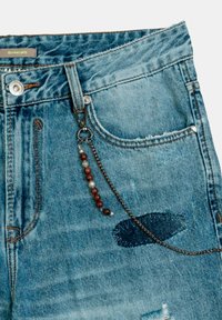 Jeans de mezclilla azul claro con bordes deshilachados, mostrando una cadena con cuentas de madera y acentos metálicos sujetada al bolsillo delantero.