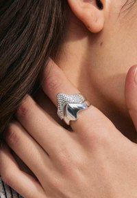 Sølv ring med bølgedesign og glat finish, prydet med indlagte rhinsten, som hviler på en hånd tæt på øret.