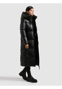 Cappotto lungo nero imbottito con un esterno lucido e trapuntato, cappuccio oversize e tasche laterali, abbinato a stivali neri chunky.
