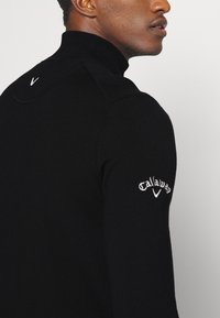 Sweater de malha preta com gola alta, apresentando textura canelada, design ajustado e logótipo "Callaway" bordado em branco na manga esquerda.