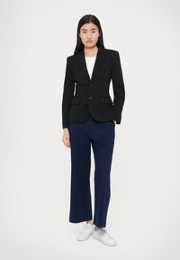 Blazer preto com dois botões, corte ajustado e lapelas com entalhe; combinado com calças largas azul-marinho e ténis brancos. Linhas limpas e design estruturado.
