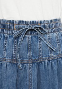 Džinsa svārki ar elastīgu jostu un regulējamu auklu. Zilas krāsas ar redzamām šuvēm un krokojumiem, piešķirot papildus tekstūru un detaļas.