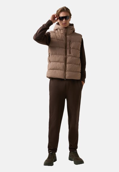 Braune Pufferweste mit Kapuze und Reißverschlusstasche, kombiniert mit einem braunen Trainingsanzug. Das Model trägt Sonnenbrillen und robuste braune Schuhe.