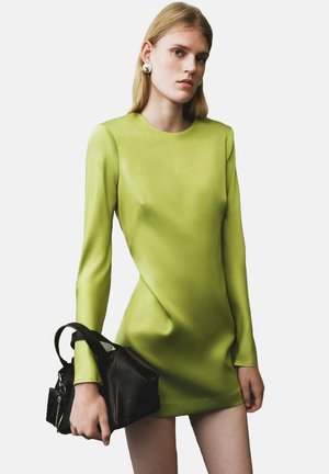 Minidress verde a maniche lunghe con una silhouette elegante e una texture liscia. Abbinato a una piccola borsa nera, dotata di un manico corto.