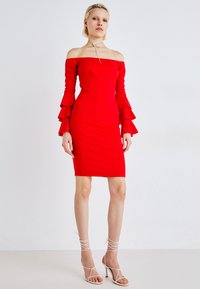 Rød off-shoulder kjole med flagrende volanger, tettsittende silhuett og knelengde. Brukt med hvite høyhælte sandaler med stropper.