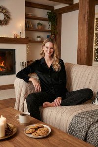 Vrouw glimlachend in zwarte pyjama, zittend met gekruiste benen op een beige bank vlakbij een salontafel met koekjes en koffie bij een open haard.