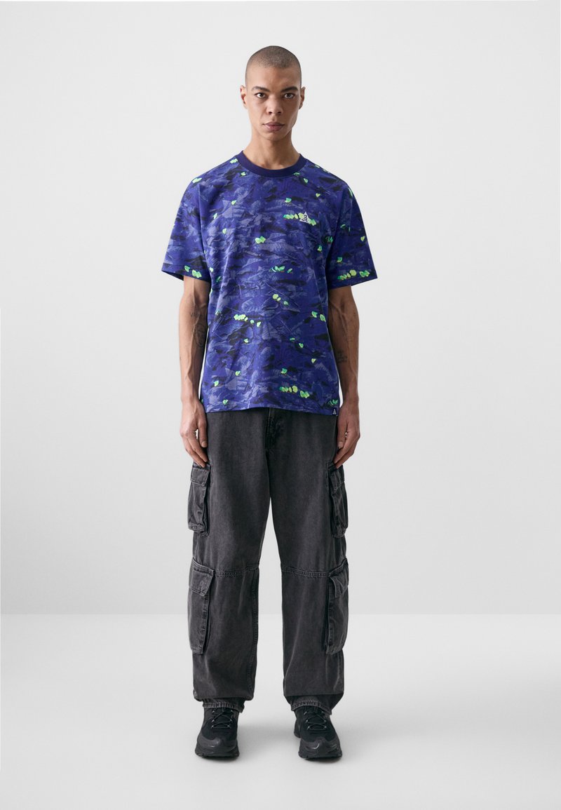 Purple short-sleeve shirt with a blue and green abstract design, paired with dark cargo pants and black sneakers.