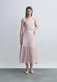 Žena v bezrukávových midi šatách svetloružovej farby s vzorom, so štvorcovým výstrihom, v bielych špicatých topánkach, stojaca pred jednoduchým pozadím.