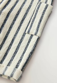 Kreemjatine puuvillakangas, millel on tumesinised vertikaalsed triibud, koos volditud manseti ja silmapaistva taskuga lisadetailideks.