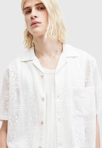 Hvid kortærmet skjorte med knapper og et blomstret mønster, som har en brystlomme og træknapper. Blødt, let stof.