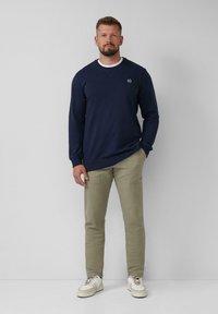 Man standing against white wall wearing navy sweatshirt, beige pants, and white sneakers, with one hand in pocket.