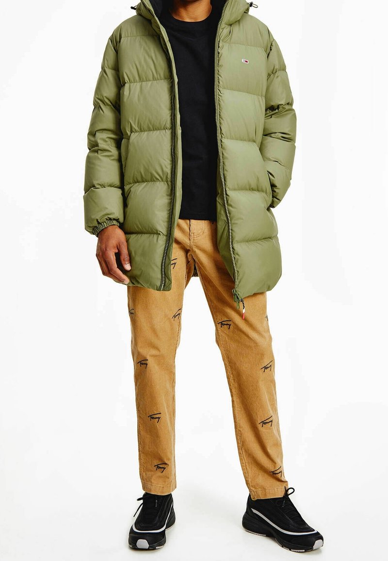 Grön puffjacka med huva, svart skjorta under, och beige byxor med svarta logomönster. Svarta sneakers fulländar outfiten.