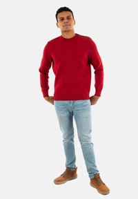 Maglione rosso lavorato a maglia con orlo a coste e bottoni sulle spalle, abbinato a jeans azzurri chiari e stivali marroni con lacci, esposto su uno sfondo bianco.