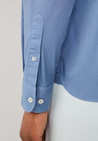 Camisa de manga larga azul claro con puño puntiagudo, dos botones blancos y costuras visibles, fabricada en un tejido suave.