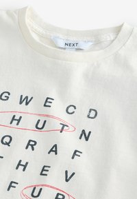Cremefarbiges T-Shirt mit schwarzem Text, der in einem Raster angeordnet ist, wobei einige Buchstaben rot umrandet sind. Rundhalsausschnitt, aus weichem Baumwollmaterial.