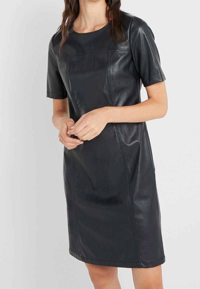 Femme portant une robe courte moulante en faux cuir noir à manches courtes avec des détails de coutures visibles, debout les mains jointes devant un fond uni.