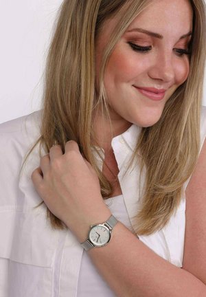 Femme souriante aux longs cheveux blonds portant un haut blanc sans manches et une montre argentée au poignet gauche.