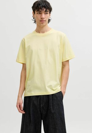 Jeune homme aux cheveux courts et foncés portant un t-shirt à manches courtes jaune pâle et un pantalon noir ample, debout avec une main dans la poche.
