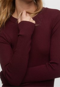 Burgundské žebrované triko s dlouhými rukávy, kulatým výstřihem a přiléhavými manžetami, vyrobené z měkké texturované tkaniny. Pohled na horní část těla ukazující ruku a paži.