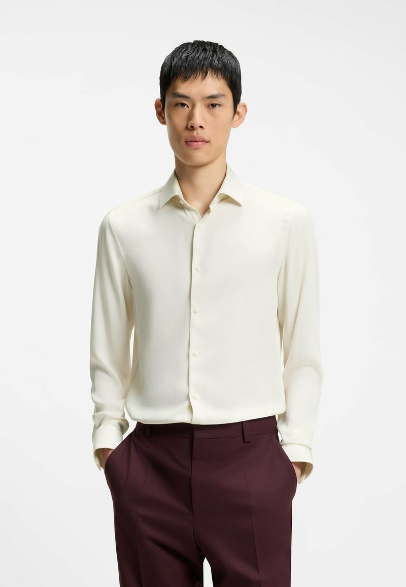 Jeune homme portant une chemise crème à manche longue boutonnée, rentrée dans un pantalon bordeaux foncé, debout les mains dans les poches sur fond blanc.