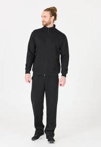Zwarte ritsjack met hoge kraag, gecombineerd met bijpassende zwarte broek. De stof lijkt zacht met een gladde textuur. Heeft zijzakken.