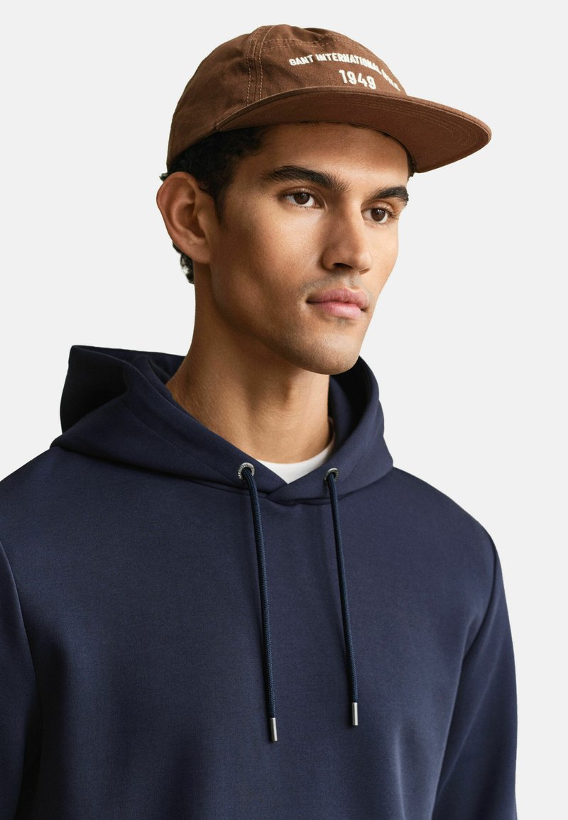 Junger Mann trägt eine braune Mütze mit weißem Text und einen marineblauen Kapuzenpullover mit Kordelzug, schaut frontal auf einen schlichten hellen Hintergrund.
