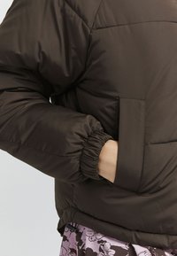 Hnedá pufová bunda s elastickými manžetami, prešívaným dizajnom a bočnou vreckom. Materiál vyzerá hladko a mierne leskle.
