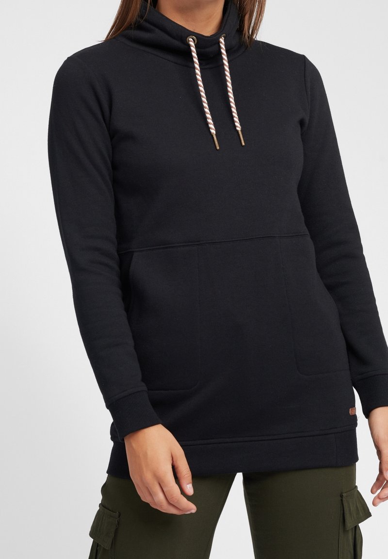 Femme portant un sweat à capuche noir à manches longues avec poches avant et cordons rayés blanc-marron, associé à un pantalon cargo vert olive.