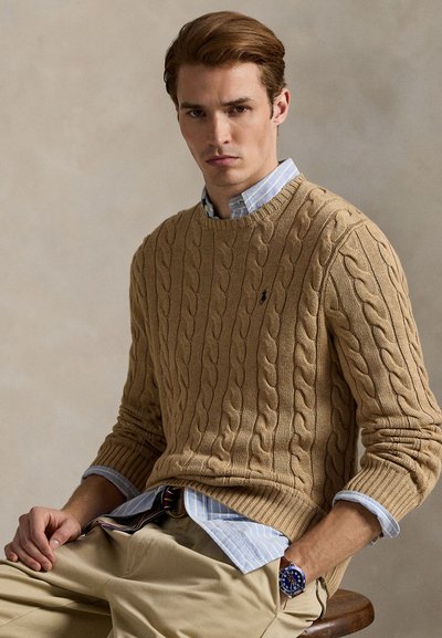 Man draagt een beige kabelgebreide trui over een blauw gestreept overhemd en beige broek, zittend op een houten kruk tegen een neutrale achtergrond.