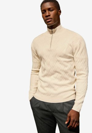 Suéter de punto beige con cuello alto y cremallera de cuarto, con una textura de patrón de diamante. Combinado con pantalones oscuros grises.